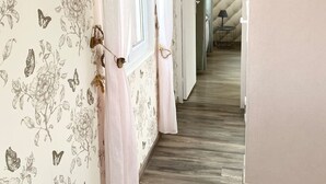 Ein Flur mit floralem Wandpapier, Holzboden und Blick in einen anderen Raum mit einer Lampe.