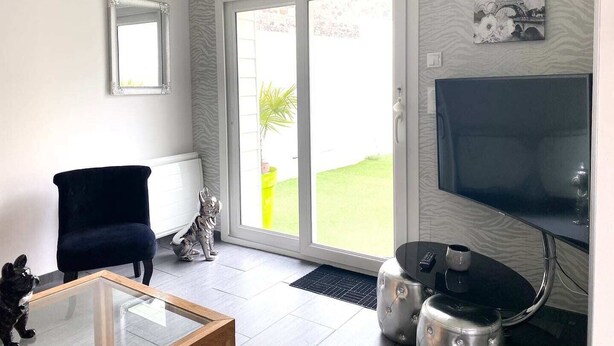 Ein Wohnzimmer mit einem schwarzen Sessel, einem Glastisch, einer silbernen Hundestatue, einem Fernseher und einer Tür, die nach draußen führt.