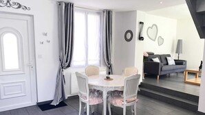 Ein Essbereich mit weißem Tisch und Stühlen, ein Fenster mit Vorhängen sowie ein Wohnbereich mit einer Couch und einer Lampe.