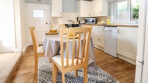 A modern kitchen with white cabinets, a dining table with chairs, and a window with a view of greenery. - Cosy Cottage (Bexhill-on-Sea)