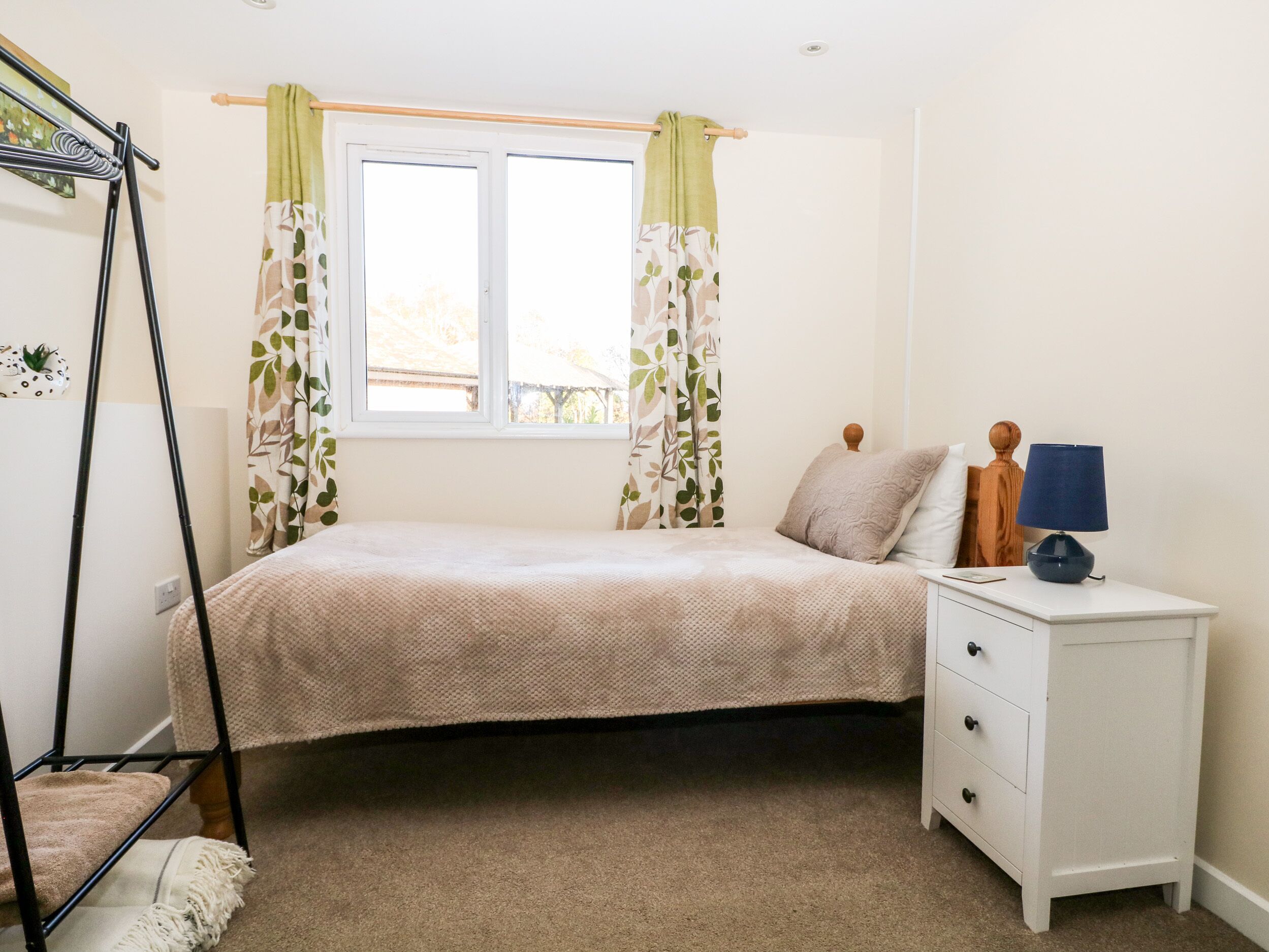 A bedroom with a bed, a nightstand, a lamp, a window with curtains, and a wall-mounted rack.