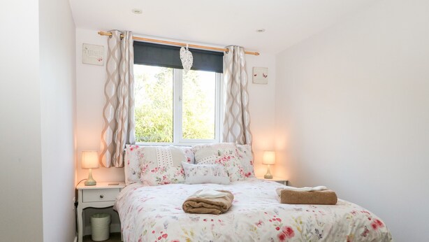 A bedroom with a bed, two bedside tables, a window with curtains, and a small bedside lamp on each table. - Cosy Cottage (Bexhill-on-Sea)