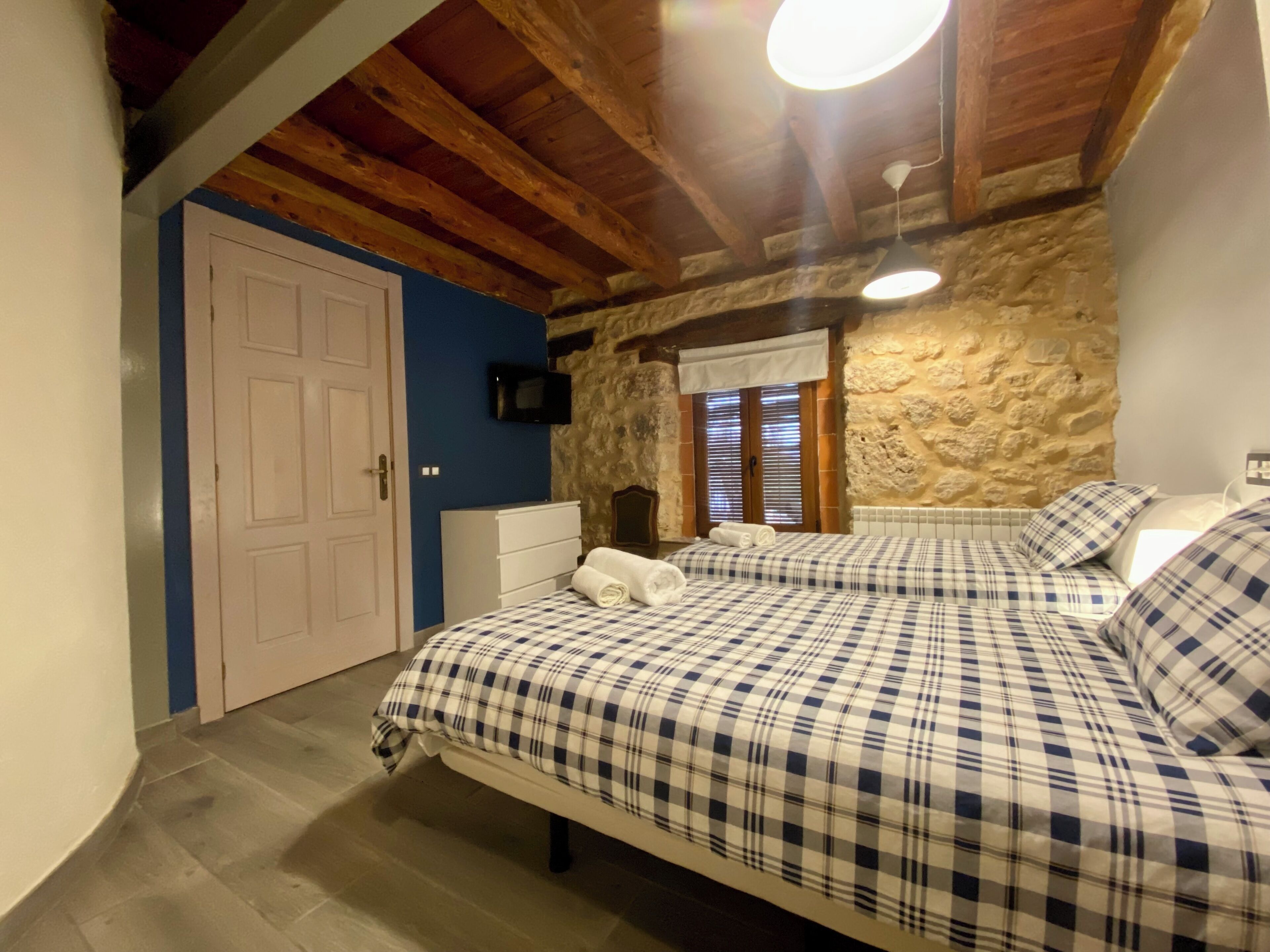 Un dormitorio con colcha a cuadros, cómoda blanca, televisor en la pared y pared de piedra.