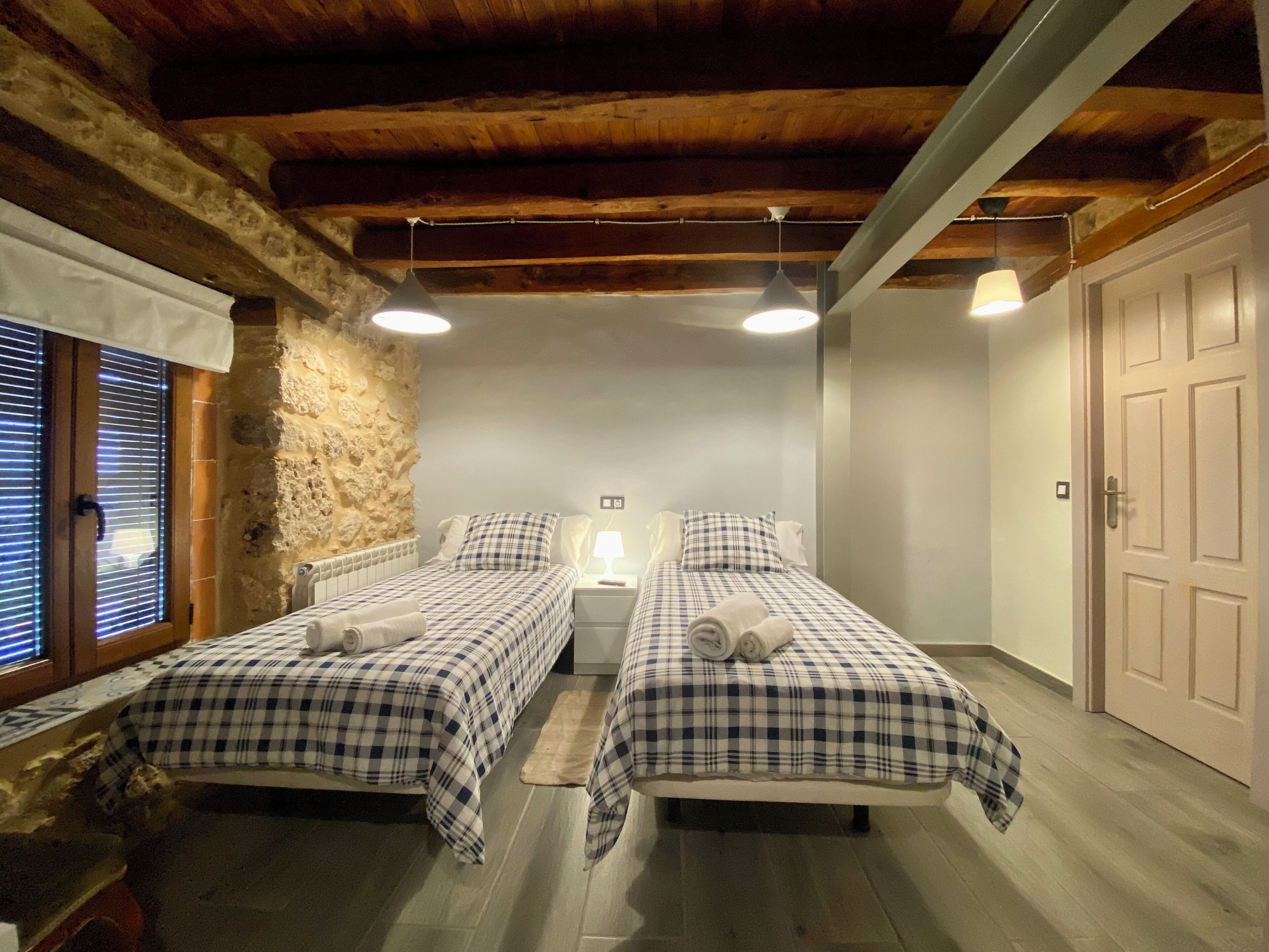 Una habitación con dos camas, cada una con colcha a cuadros, una mesita de noche con lámpara y una pared de piedra.