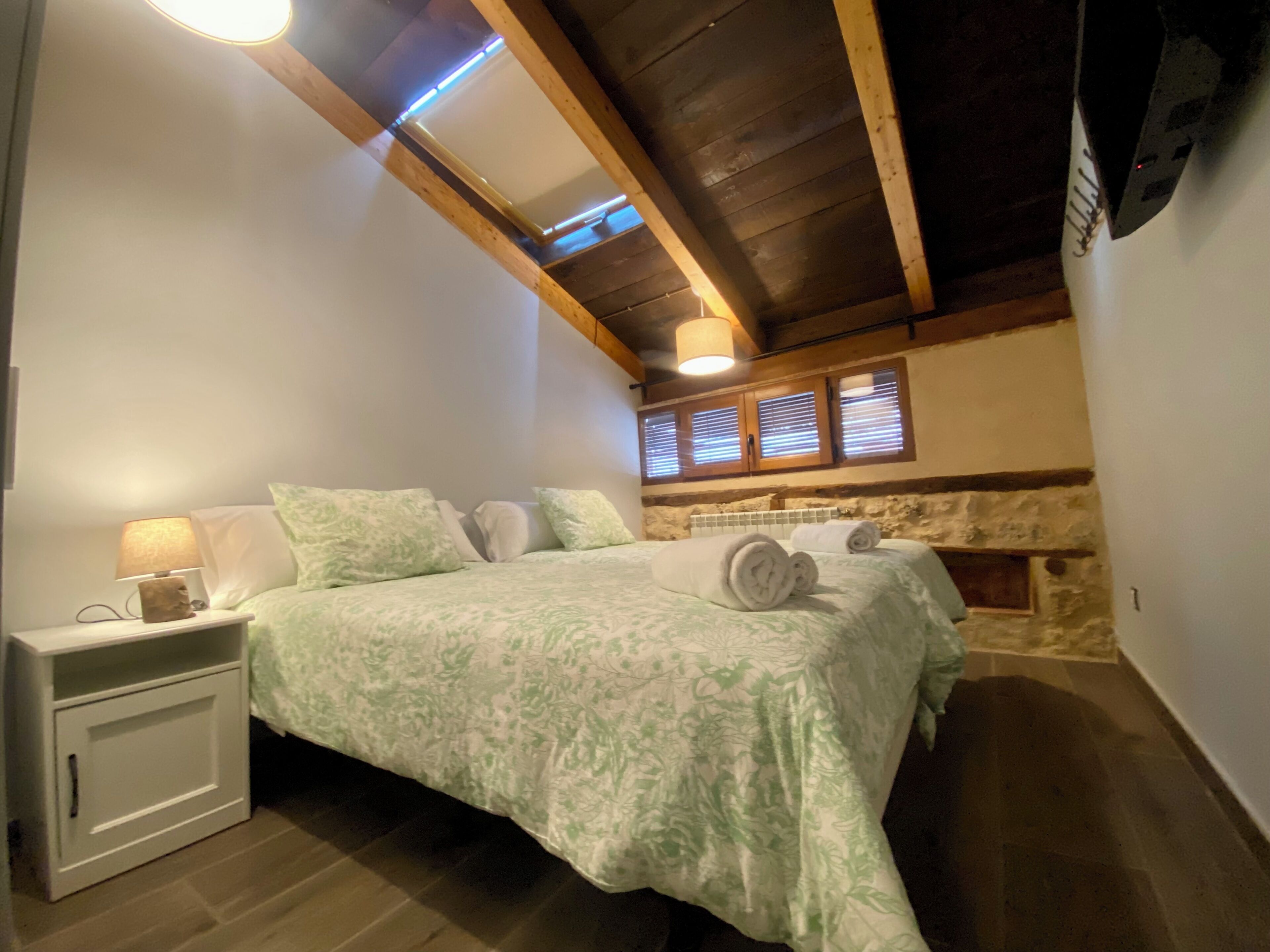 Un dormitorio con cama, mesita de noche, lámpara, pared de piedra y techo de madera.