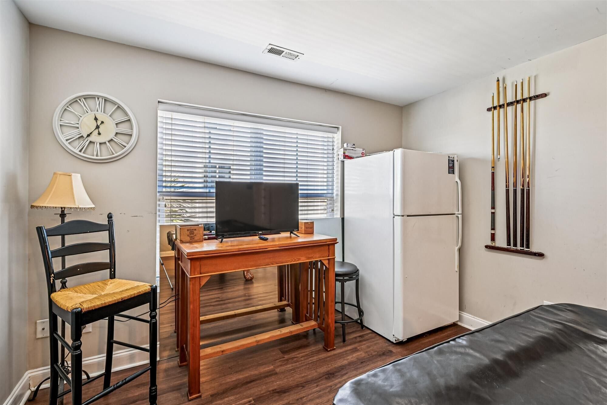 A compact room with a wooden desk, a television, a refrigerator, a bed, a chair, a clock, and a window with blinds.
