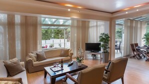 A spacious living room with a sofa, coffee table, and chairs, a television on a stand, and a balcony with plants and a dining area. - A Private Haven: Hera with Secluded Pool and Garden (Kassandra)