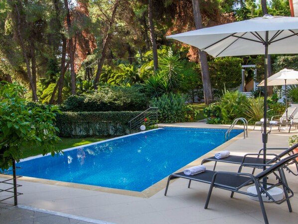 Une piscine rectangulaire dont les bords sont recouverts de carreaux, entourée d’une terrasse également carrelée. On y trouve des chaises longues en métal et un parasol blanc.