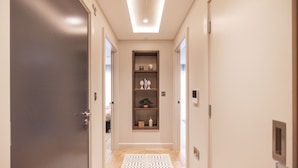 A hallway with a dark door on the left, a light-colored wall, a ceiling light, and a shelf with various items.