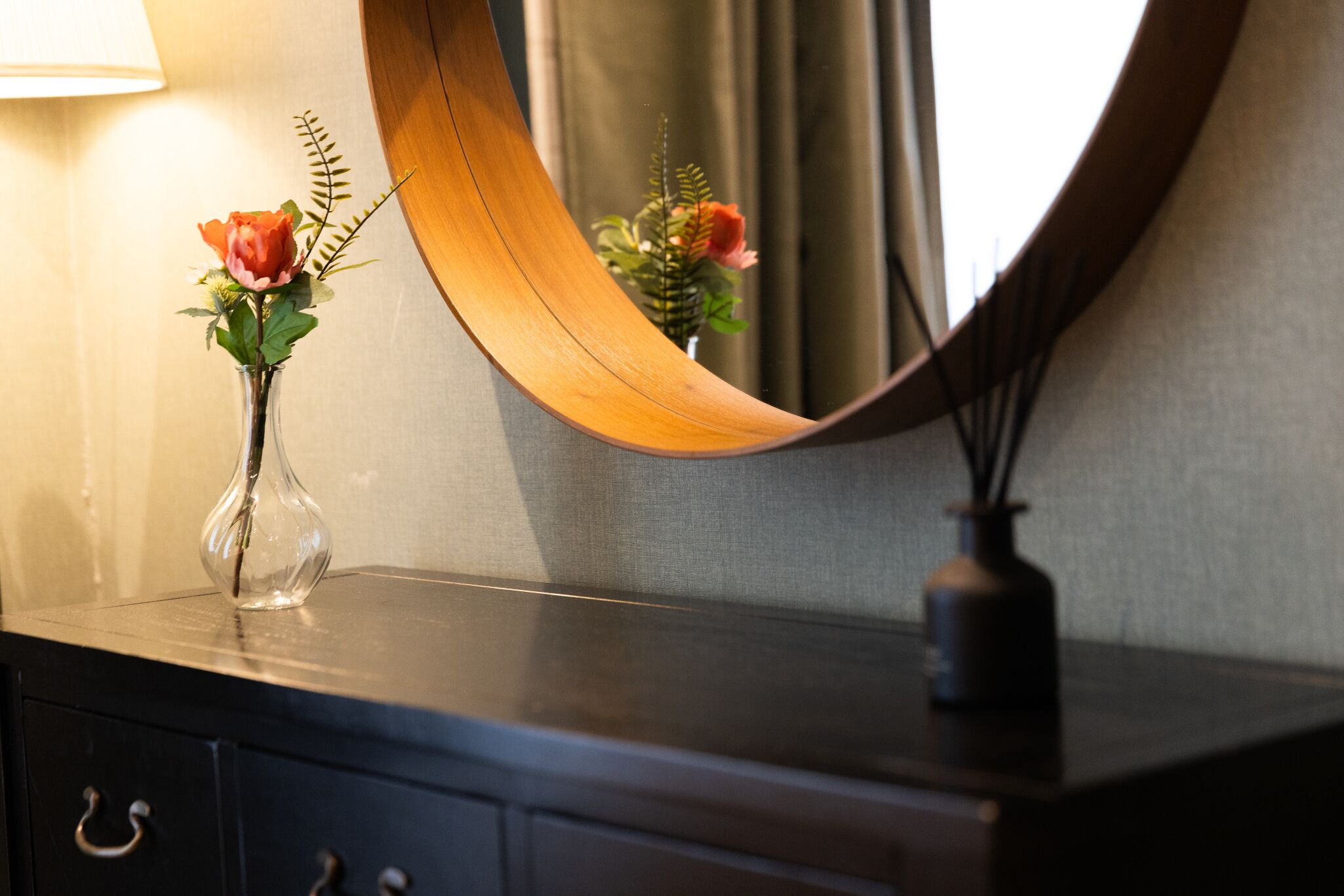 Uno specchio curvo in legno che riflette un vaso con fiori e una pianta, posizionato su una cassettiera di legno scuro con una lampada sullo sfondo.