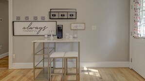 A room with a white wall, a framed sign saying 'always thankful,' a coffee machine, and a small table with two chairs.
