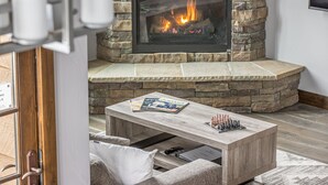 A stone fireplace with a fire, a wooden coffee table with magazines and chess pieces, and a cushioned chair.