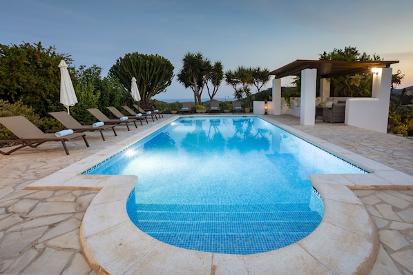 Una piscina con sillas de descanso y un área de estar cubierta, rodeada de vegetación y vistas al paisaje.
