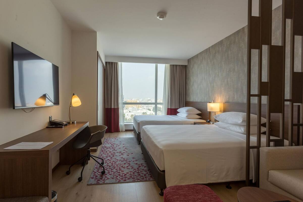 A hotel room with a desk, two beds, a chair, a TV, and a view of the city.