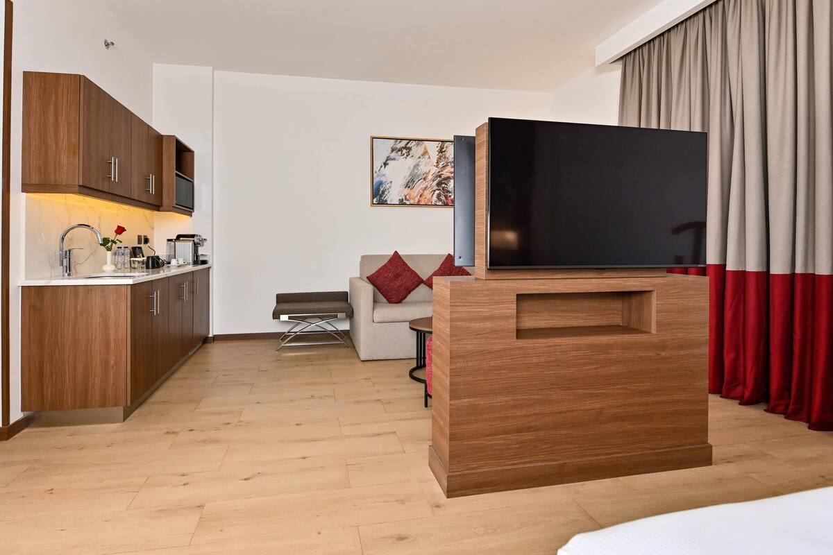 A modern hotel room with a wooden kitchenette, a flat-screen TV, a sofa with red cushions, and a large window with curtains.
