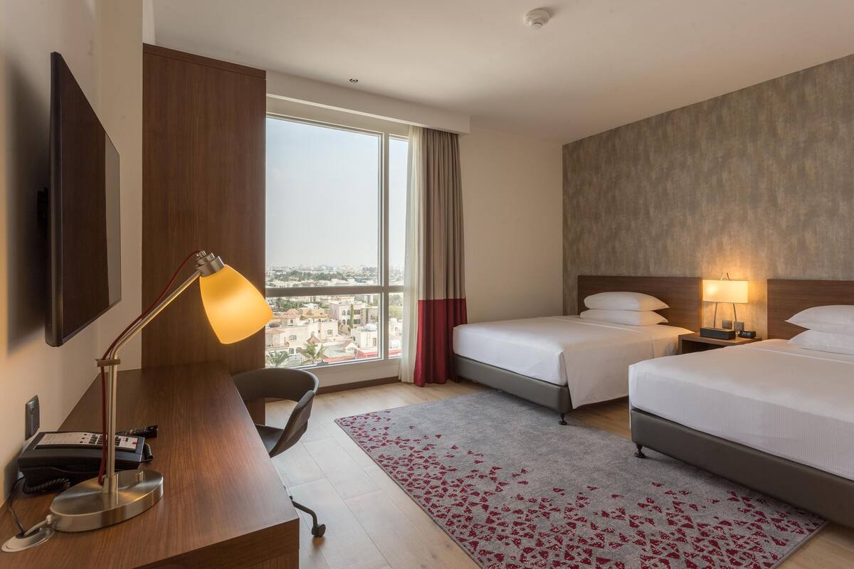 A hotel room with two beds, a desk with a lamp, a chair, a desk phone, and a view of the city through large windows.