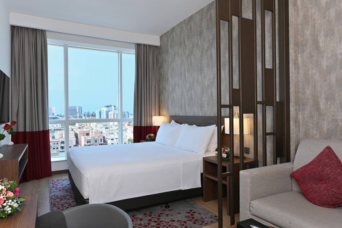 A hotel room with a large bed, a sofa, a desk with a lamp, and a view of the city through large windows.