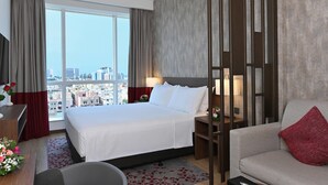 Habitación de hotel con una cama grande, un sofá, un escritorio con lámpara y vistas de la ciudad a través de amplios ventanales.