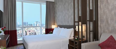 Ein Hotelzimmer mit einem großen Bett, einer Sitzecke, einem Schreibtisch mit Lampe und Blick auf die Stadt durch große Fenster.