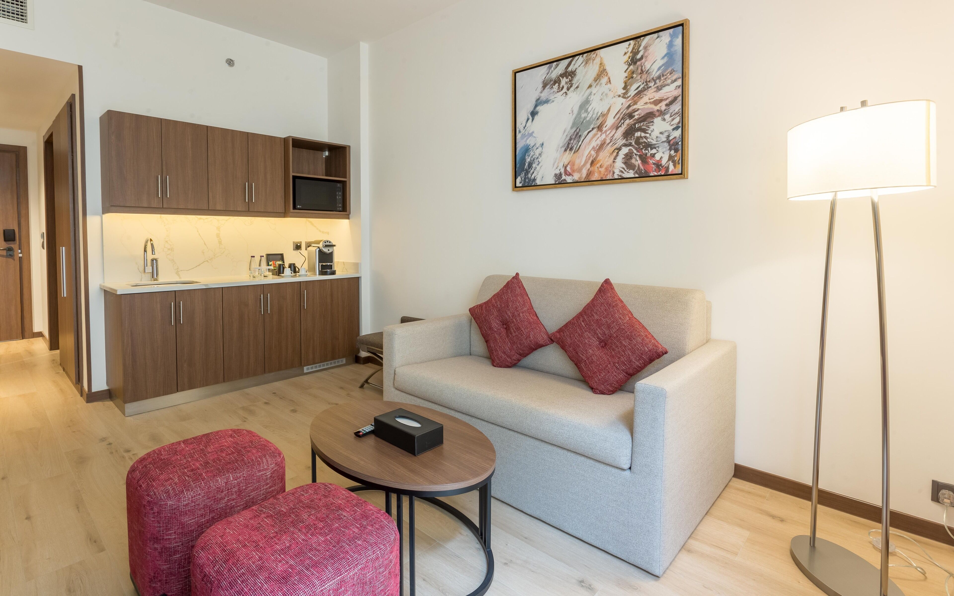 Un espace de vie compact comprenant un coin cuisine, un canapé avec des coussins rouges, une petite table ronde et un lampadaire.