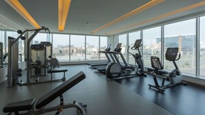 Un gimnasio moderno con cintas de correr, máquinas de pesas y una banca. Amplias ventanas ofrecen vistas de la ciudad.