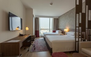 Habitación de hotel con escritorio, dos camas, una silla, televisión y vistas a la ciudad a través de amplias ventanas.