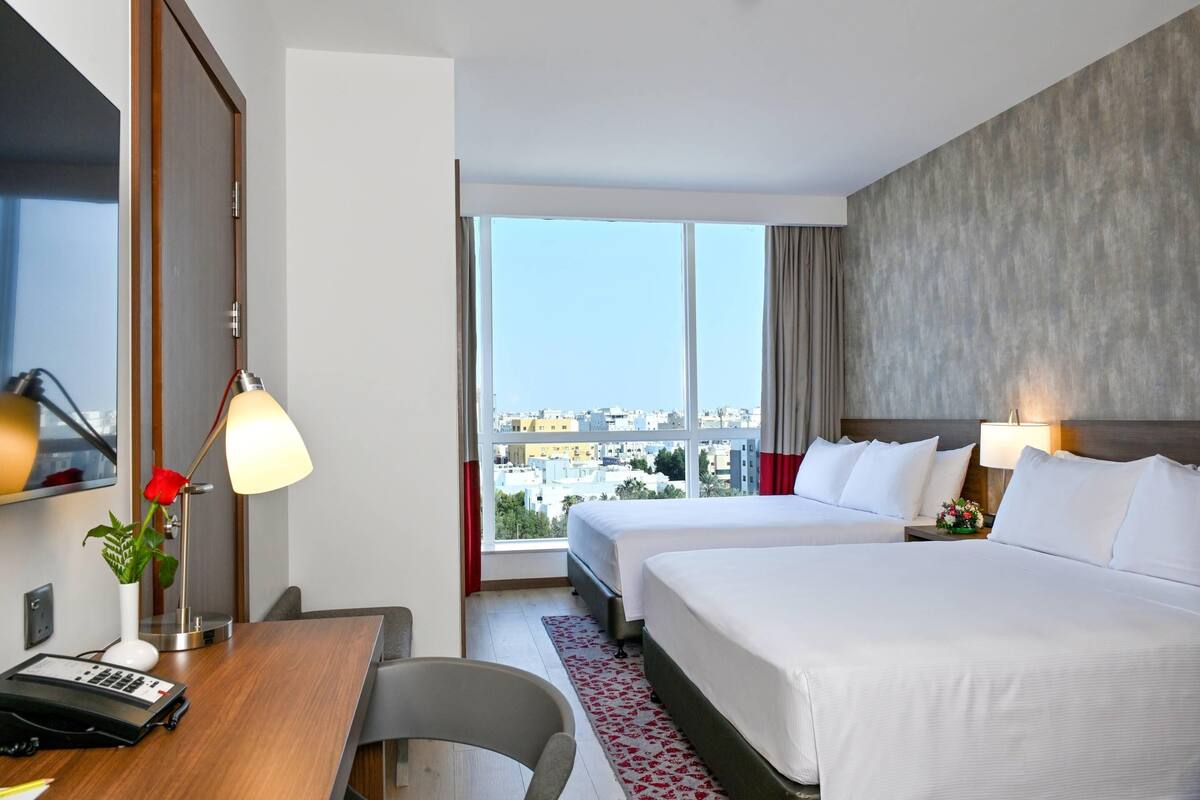 A hotel room with two beds, a desk with a lamp, a vase with a rose, and a view of the city through large windows.