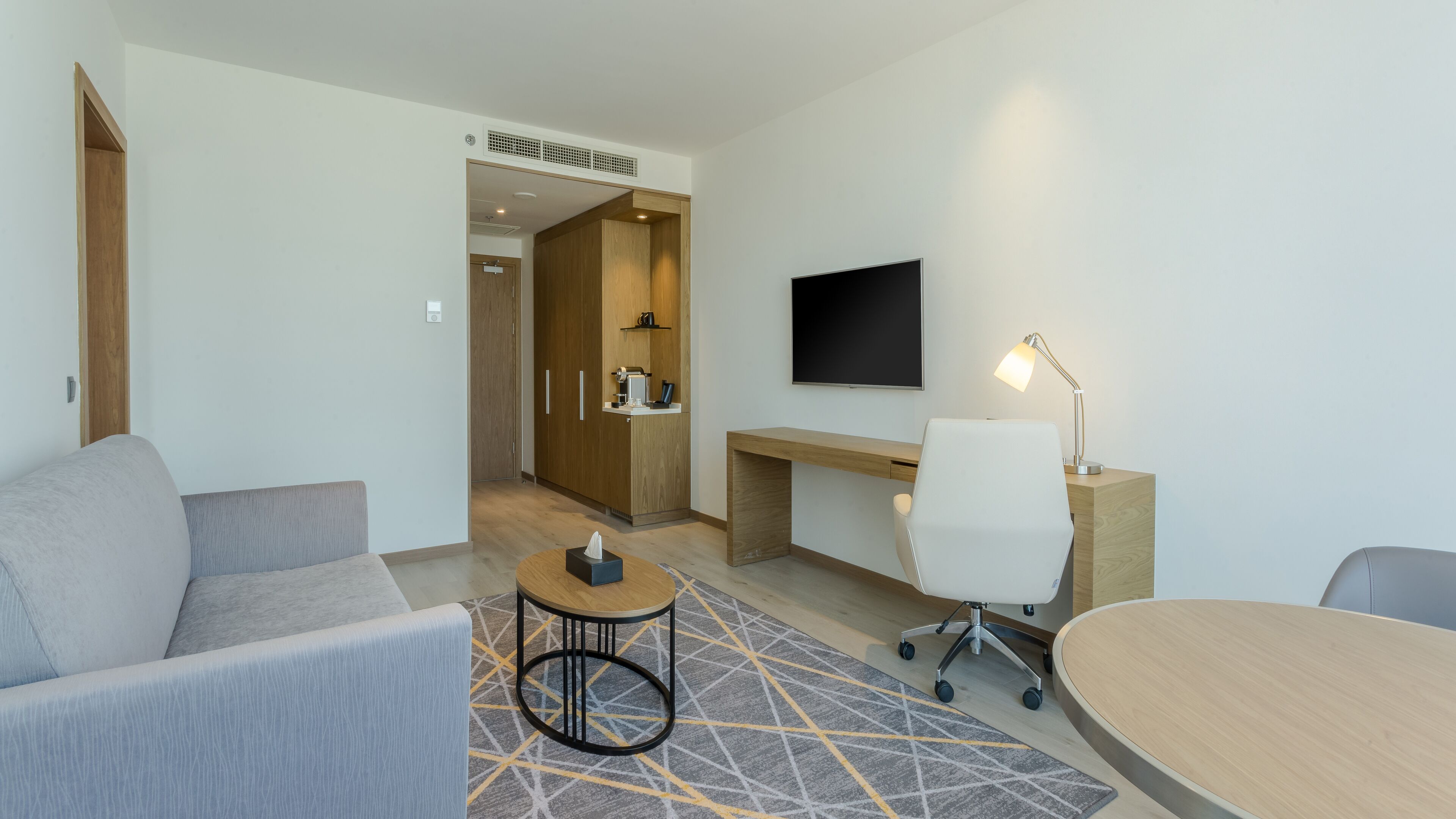 A modern hotel room with a sofa, a desk with a chair, a TV, and a small table with a lamp.