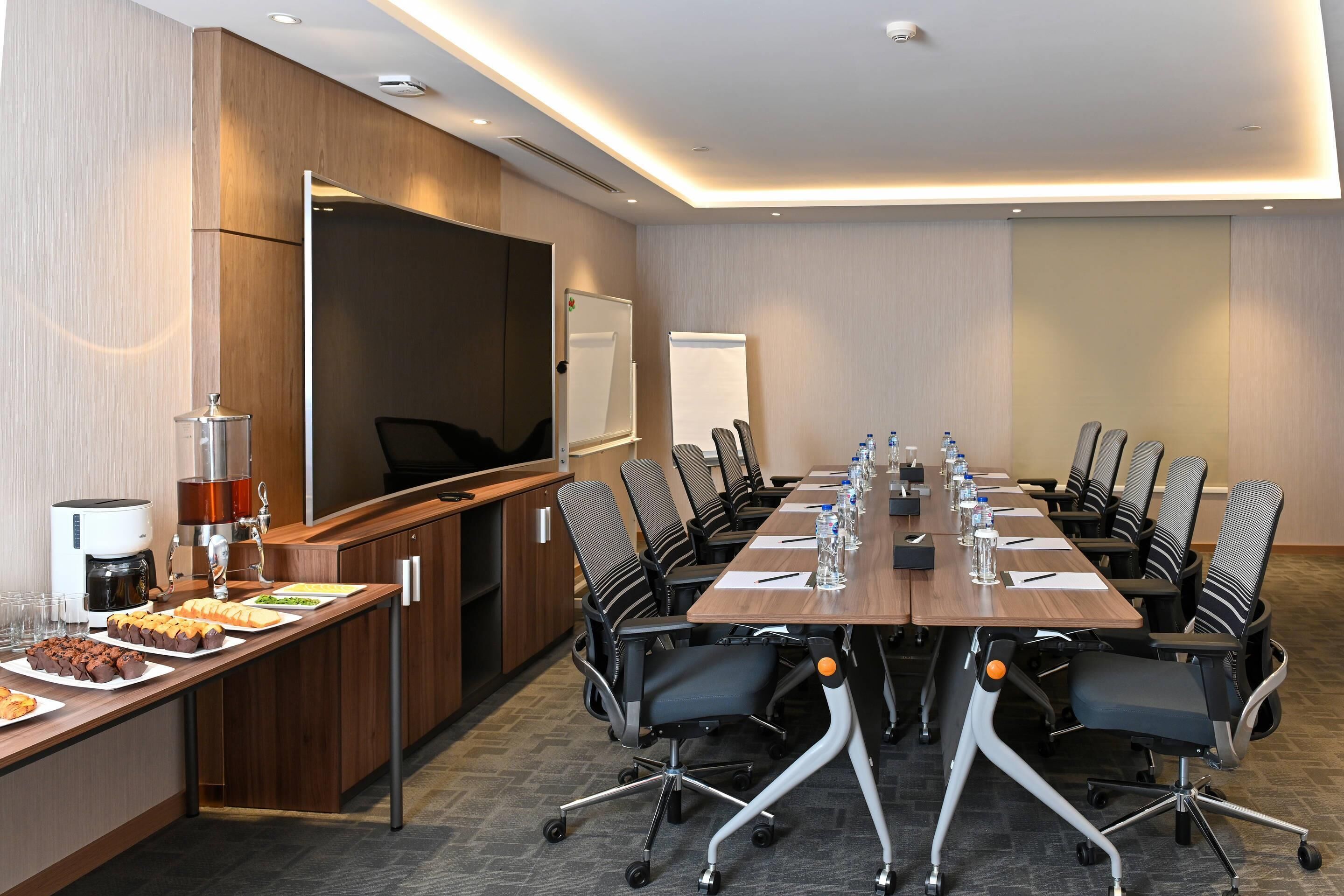 A conference room with a long table, chairs, a coffee station, and a large screen.