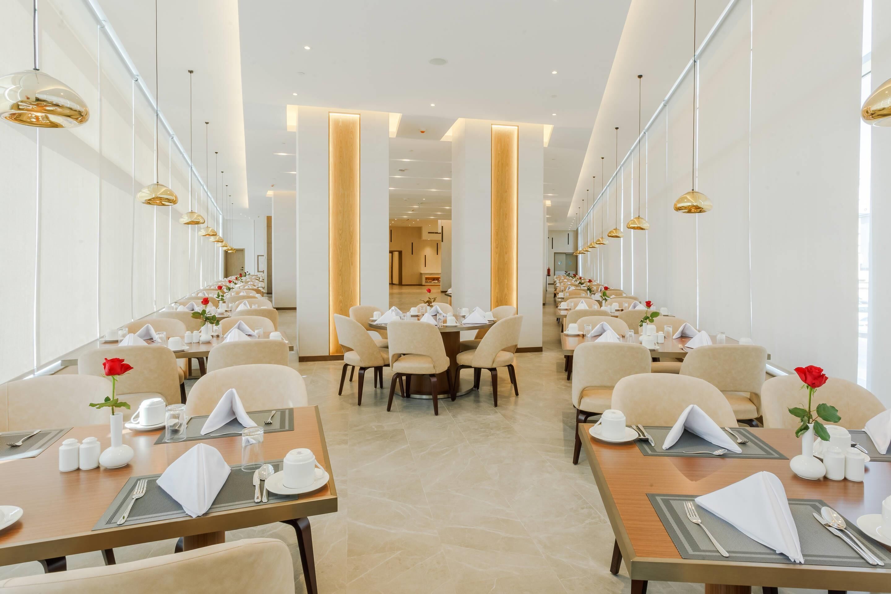 A modern hotel dining area with long tables, beige chairs, and gold pendant lighting.