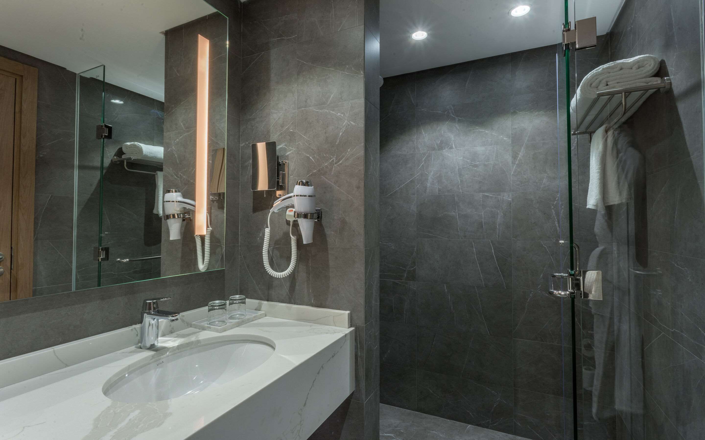 Un baño moderno con un espejo grande, un lavamanos blanco, cabina de ducha con mampara de vidrio y un perchero para toallas.