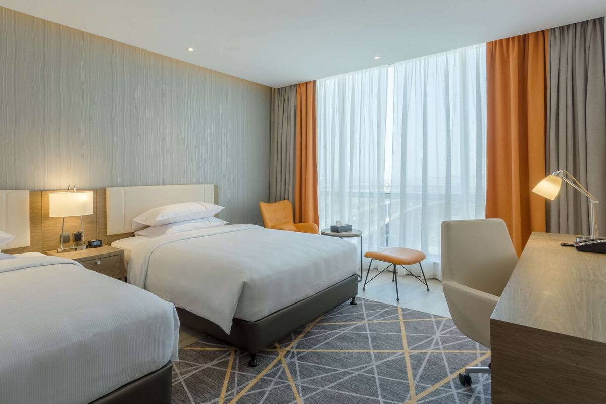 Une chambre d’hôtel avec deux lits, un bureau, une chaise et une vue sur la ville grâce à de grandes fenêtres.