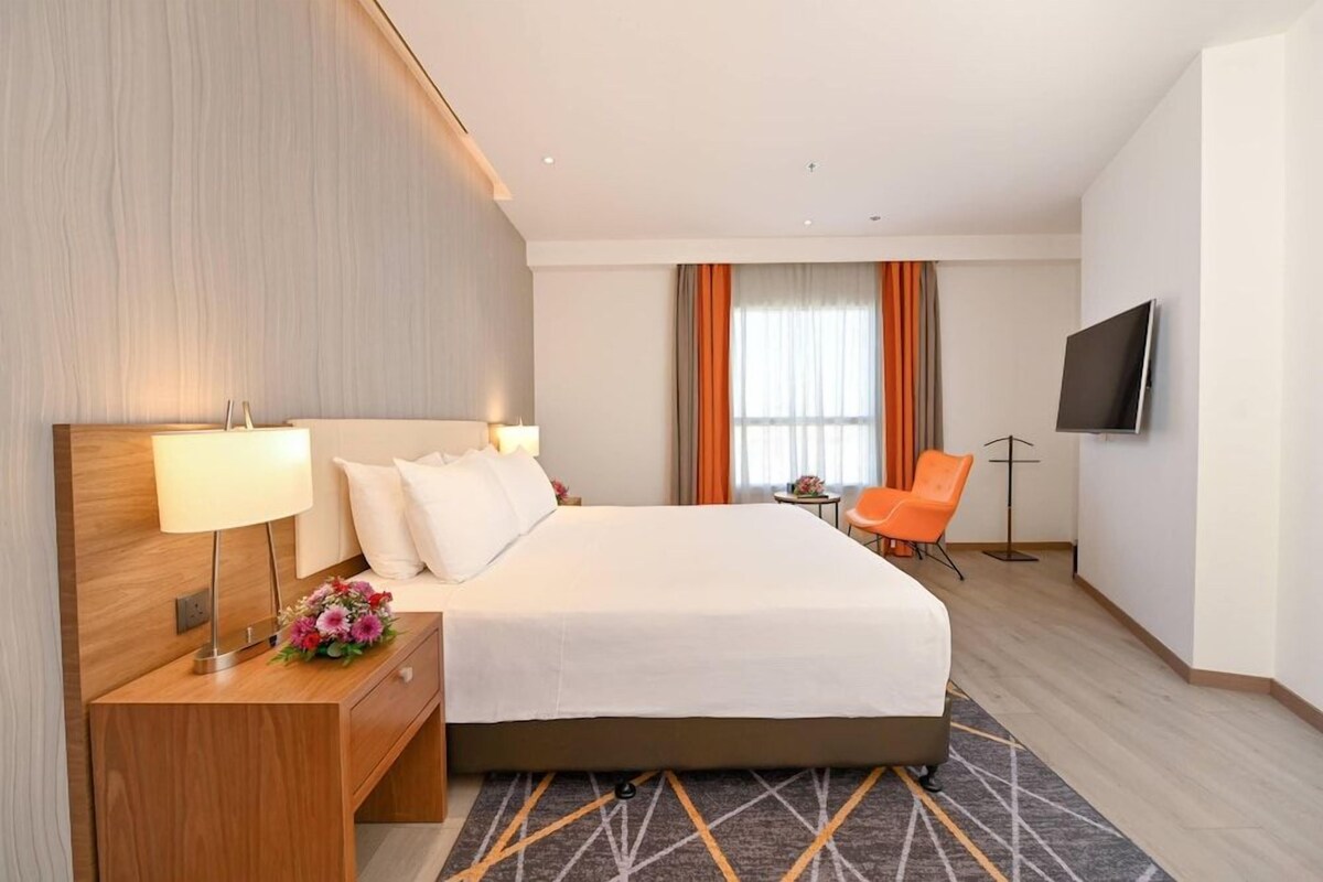 Ein Hotelzimmer mit einem großen Bett, einem Schreibtisch mit Lampe, einem orangefarbenen Sessel und einem an der Wand befestigten Fernseher.