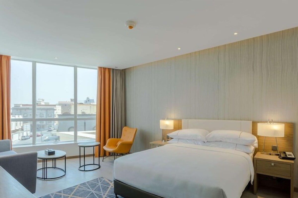 Habitación de hotel con una cama grande, dos lámparas de noche, una silla, una mesita y vistas a la ciudad a través de amplias ventanas.