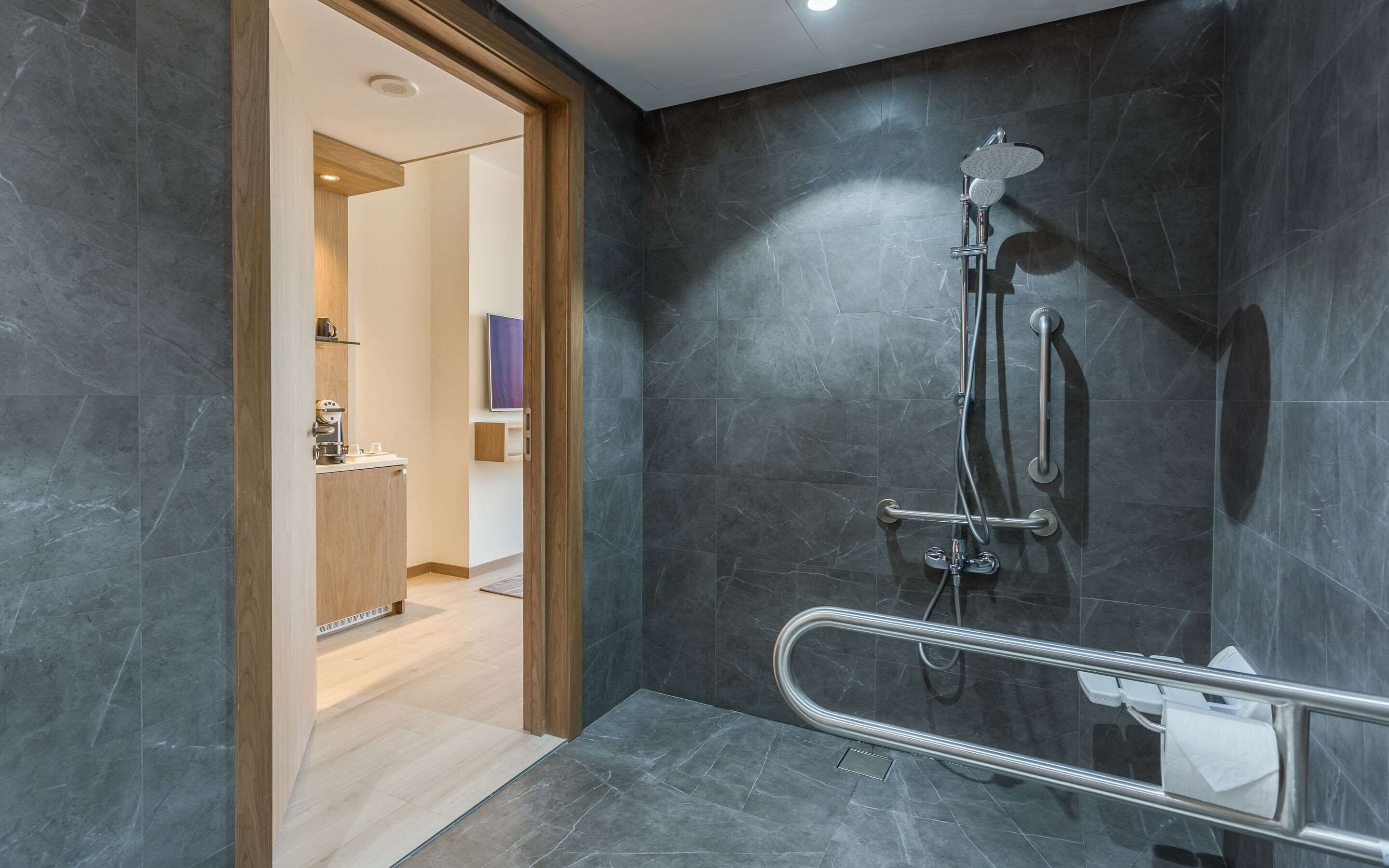 A modern bathroom with a shower, grab bar, and a wooden door leading to a bathroom vanity.