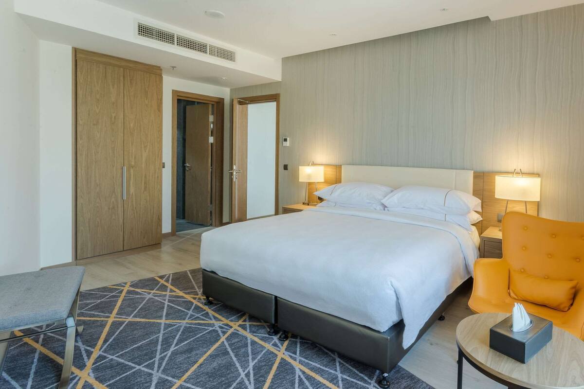 Une chambre d’hôtel avec un lit, deux lampes de chevet, une petite table avec une boîte de mouchoirs, une chaise et un tapis à motifs.
