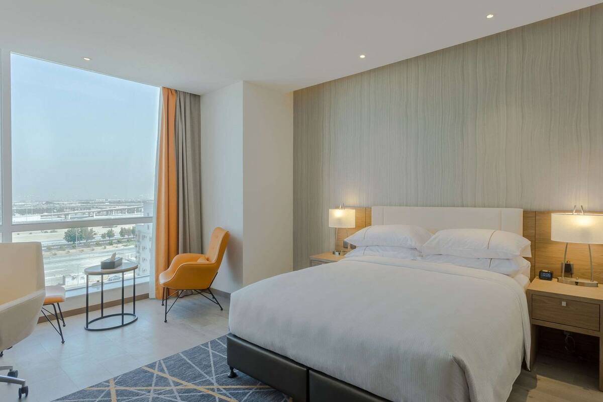 Une chambre d’hôtel avec un grand lit, deux lampes de chevet, une chaise, une petite table et une vue sur le paysage urbain.
