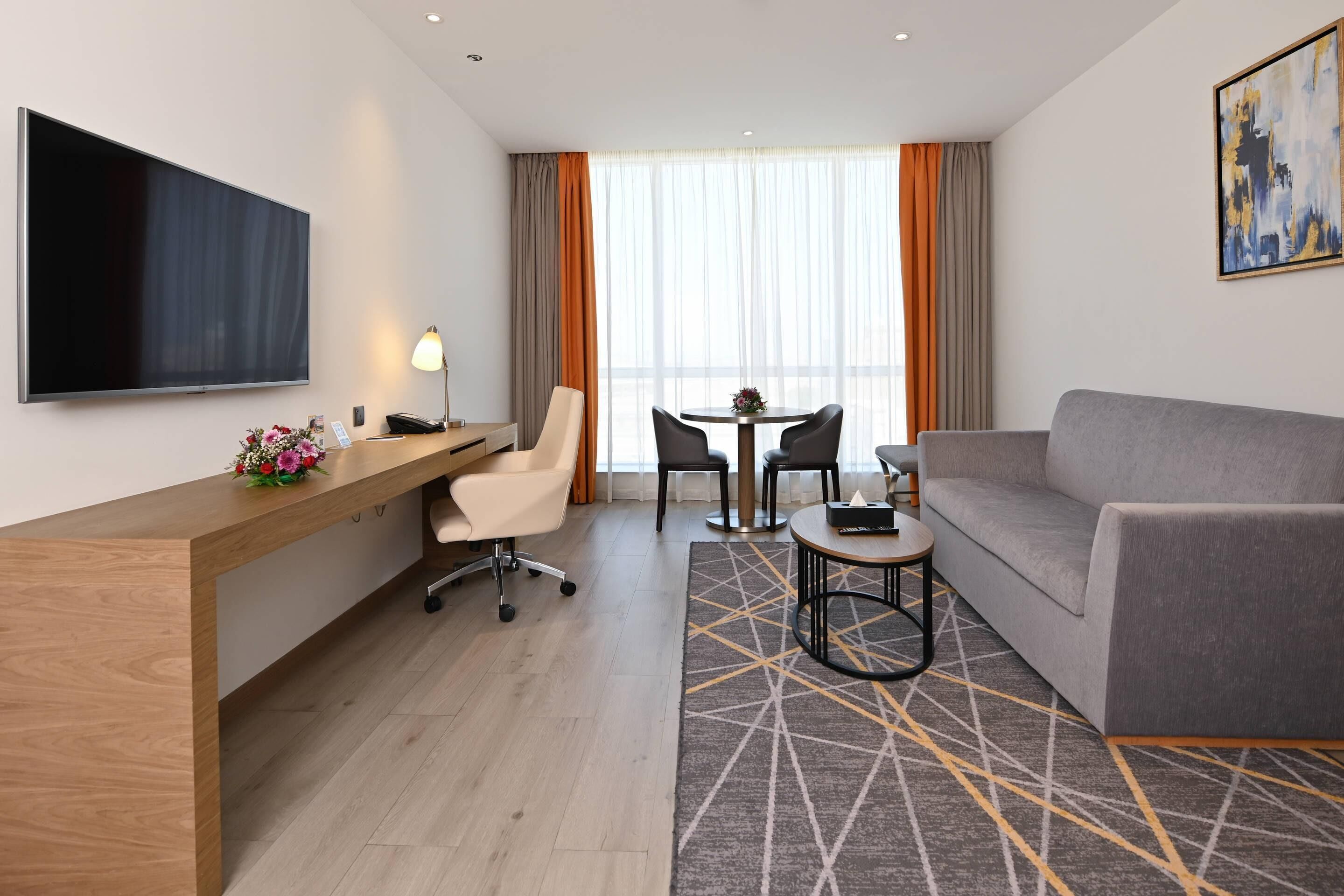 A modern hotel room with a wooden desk, a grey sofa, a TV, and a dining area with a table and chairs.