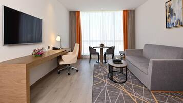 A modern hotel room with a wooden desk, a grey sofa, a TV, and a dining area with a table and chairs.