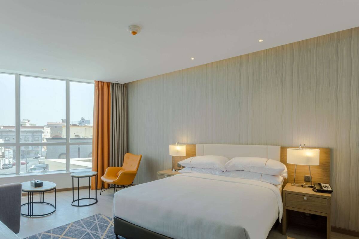 Une chambre d’hôtel avec un grand lit, deux lampes de chevet, une chaise, une petite table et une vue sur la ville grâce à de grandes fenêtres.