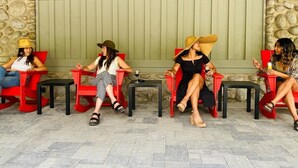 Quatre personnes sont assises sur des chaises rouges, devant des tables noires, adossées à un mur de pierre et face à une porte verte.