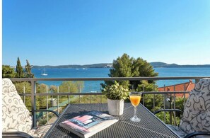 Un balcón con vista al mar, una mesa con un libro y un vaso de jugo de naranja, y una planta en maceta.