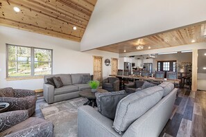 A spacious living room with a large gray sectional sofa, a round coffee table, and patterned armchairs. A wooden ceiling with recessed lighting and a large window providing a view of the outdoors. - Crown Jewel | Table Rock Lake Views | Sleeps 24 | Near Silver Dollar City (Indian Point)