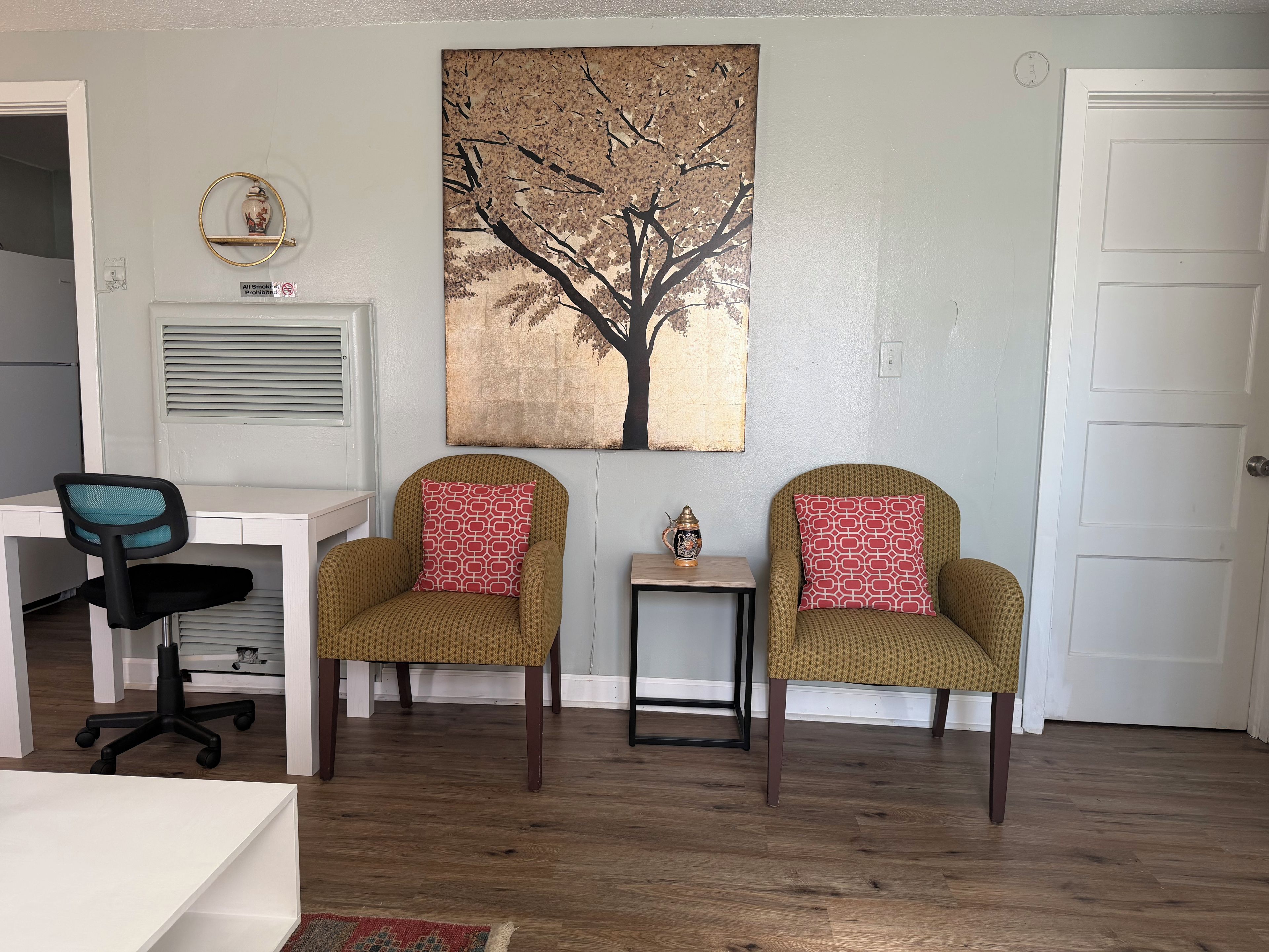 Une pièce avec un bureau blanc, deux fauteuils en osier ornés de coussins à motifs, une petite table sur laquelle est posé un objet décoratif, et un grand tableau représentant un arbre accroché au mur.