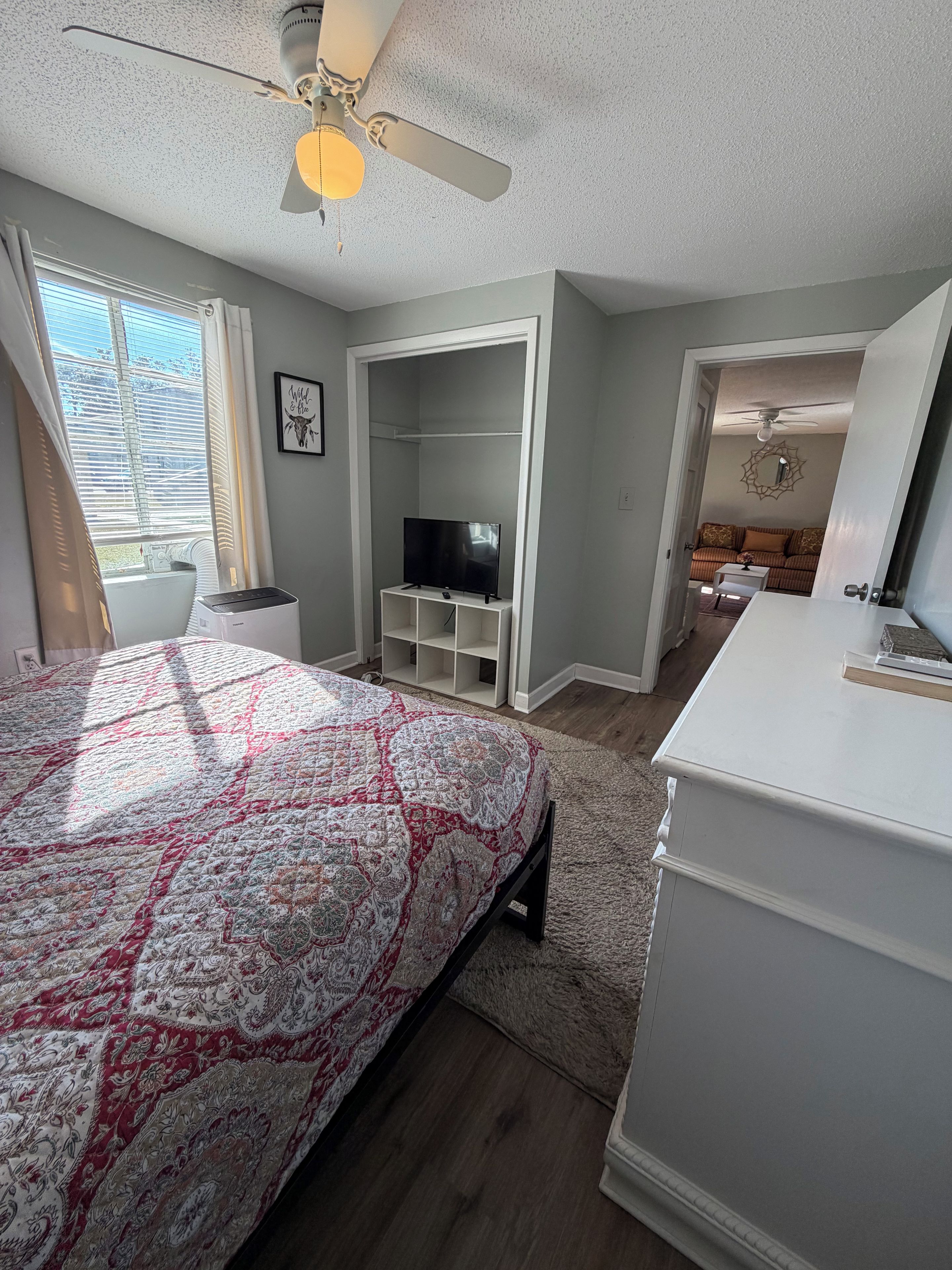 Une chambre à coucher avec un lit, un téléviseur posé sur un meuble, un ventilateur de plafond et une vue sur le salon.