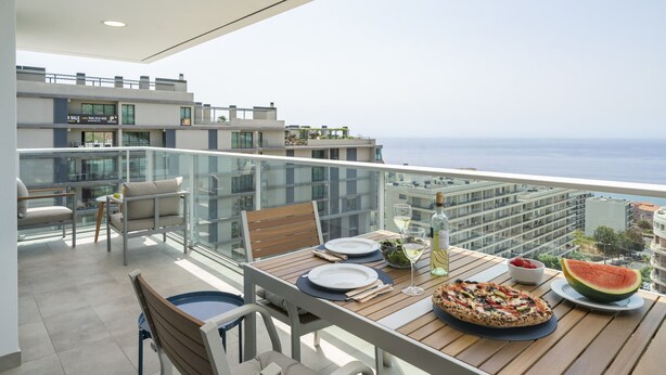 A balcony with a dining table set for two, a pizza, a watermelon slice, and a bottle of wine.