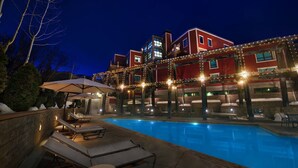 Una piscina con sillones de descanso y sombrillas, rodeada por un edificio de fachada roja y luces colgantes.