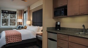 A hotel room with a bed, bedside table, lamp, and a view of trees outside.