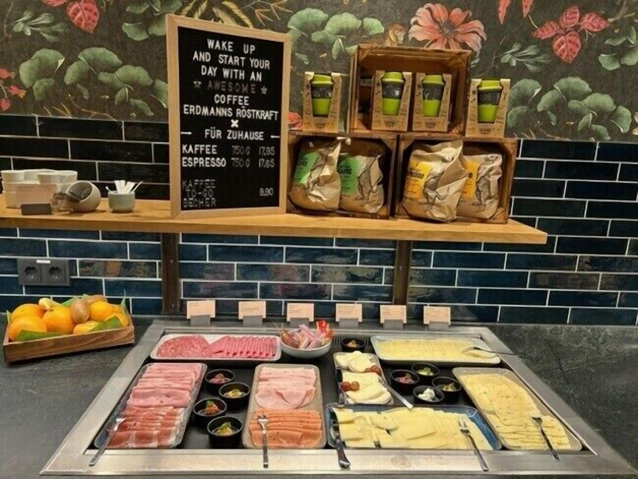 A breakfast buffet with various meats, cheeses, and fruits, along with a coffee station and a chalkboard menu.