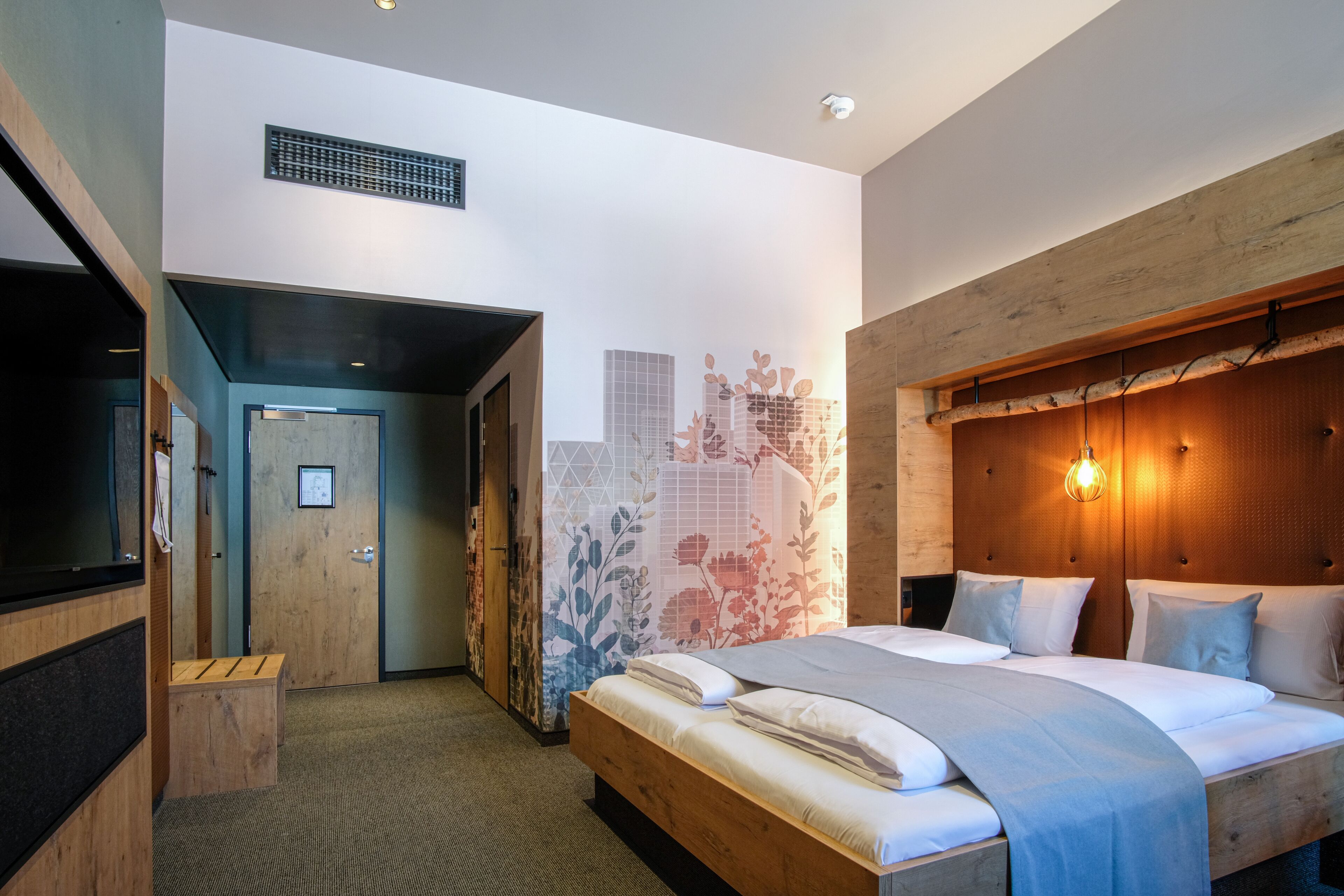A modern hotel room with a large bed, wooden headboard, and a cityscape mural on the wall.